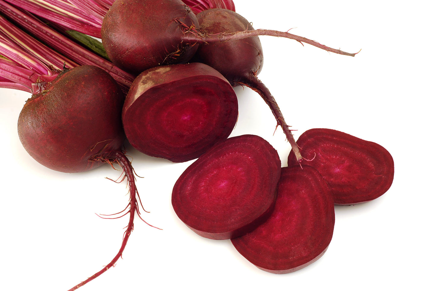 On display are 4 beets, of the variety Detroit Dark Red, laying on a white surface, surrounded with a white background. One beet is partially in front of the other 3 beets. The beet in front is cut in half, displaying the red juiciness of the beet flesh, with 3 slices laying in front. The 3 beet in the back are intact, with the stem and leaves also intact.  Prominently displayed for the 3 beets in the back are their long roots.