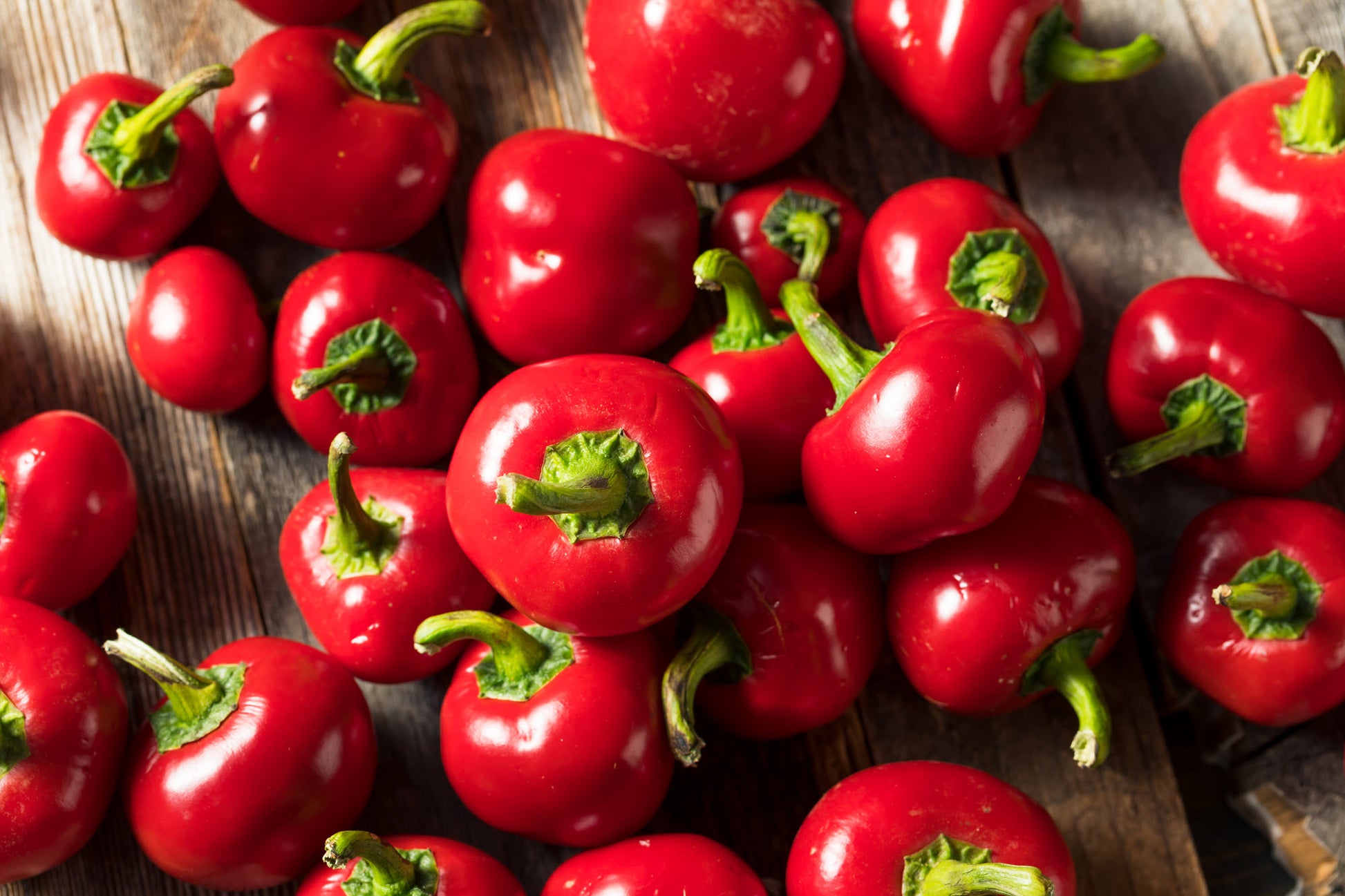 On display is a bunch of small cherry peppers, of the variety Red Cherry Sweet. Typically known as cherry bombs, these peppers are full of sweetness to enhance any salad. The peppers are stacked loosely on a weathered wooden surface. The shadows indicate a light source from the left. 