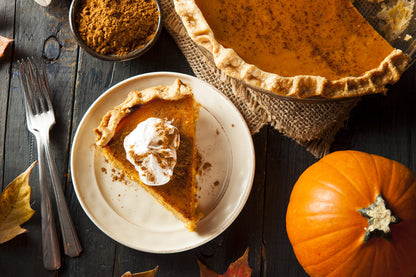 On display is a pumpkin, of the variety Sugar Pie, in lower right. In the foreground is a white dish with a slice of pumpkin pie, with whip cream, and a sprinkling of cinnamon. 2 forks appear to the left. In the upper right is the pumpkin pie, with cinnamon sprinkles, on a rope napkin. To the left of the pie dish, is a dark ceramic dish with a mound of cinnamon. Everything rests on a weathered wooden surface, with some orange fall leaves framing various points. 