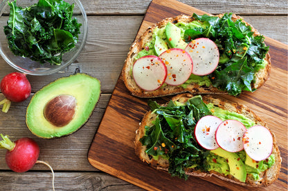 On display is a hearty serving of avocado, radishes and turnip greens, on toasted whole grain bread. The avocado is served both as a foundation of guacamole, with slices of avocado laid on top. There are 2 slices of bread topped with everything, appearing on a wooden chopping board. To the side is half of an avocado, with its seed still intact, and rest next to 2 radishes. A small glass bowl is in upper left with fresh turnip greens. 
