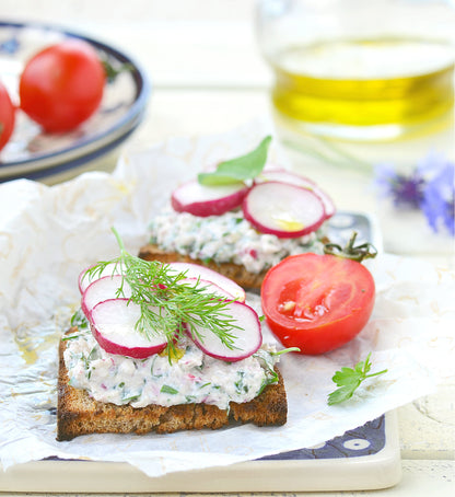 On display is a hearty serving of a dill relish, topped with radish slices and sprig of dill laid onto 2 toasted bread slices. resting on tissue paper placed on a porcelain plate, with a dark blue pattern. Everything rests on tiled surface with a white marbled look. Half of a large cherry tomato is placed next to bread. In upper left is a small blue and white porcelain dish with 2 small whole tomatoes. To upper right is a glass of olive oil. Only the bread is in focus. 