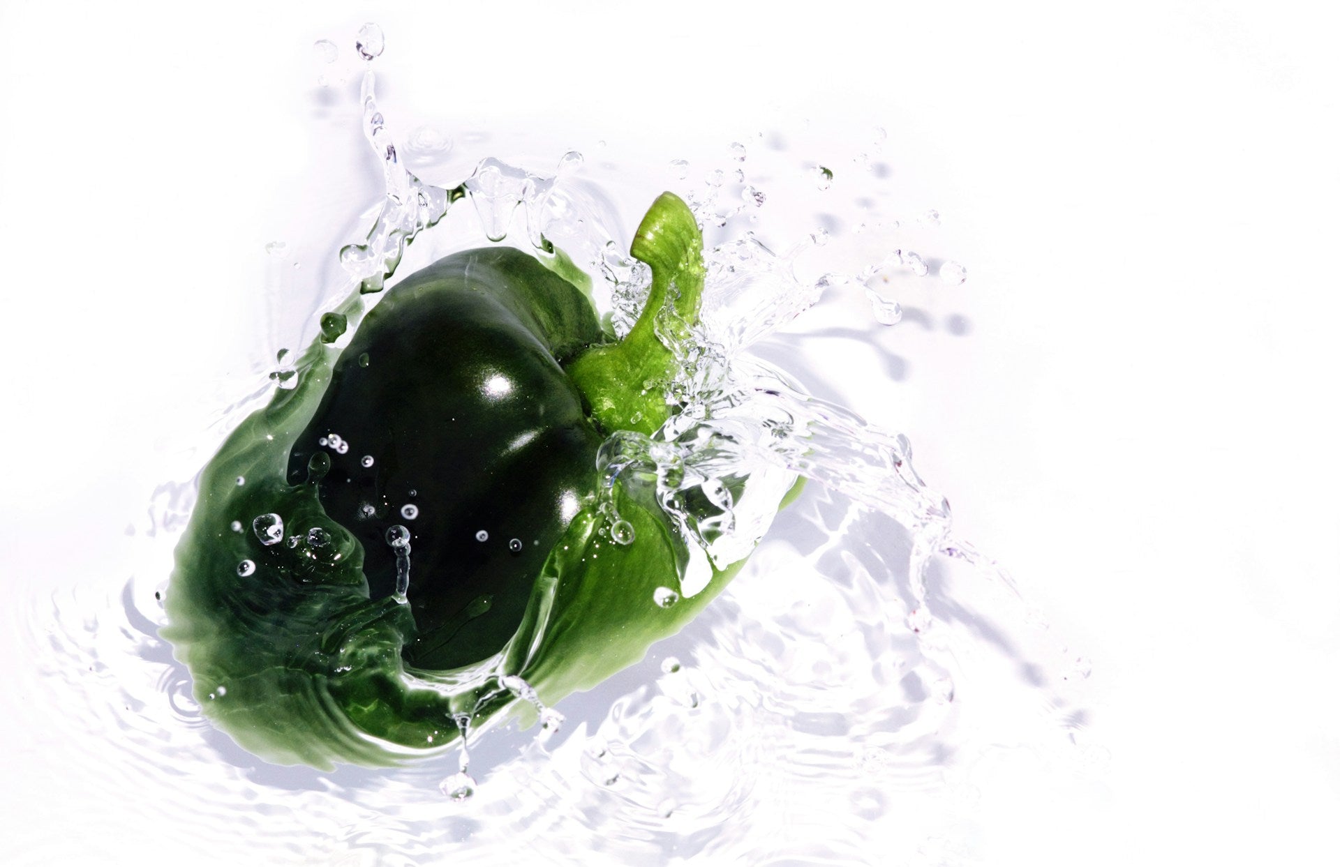 On display is a single green bell pepper, of the variety Sweet Cal Wonder 300 TMR. The pepper appears to be splashing into water, with the image capturing the immediate splash and the clear water droplets being ejected away from the splash zone. There appears to be a light source from the left, creating shadows that capture the water waves moving away. Shadows are also visible for the droplets and water that has been ejected into the air. 