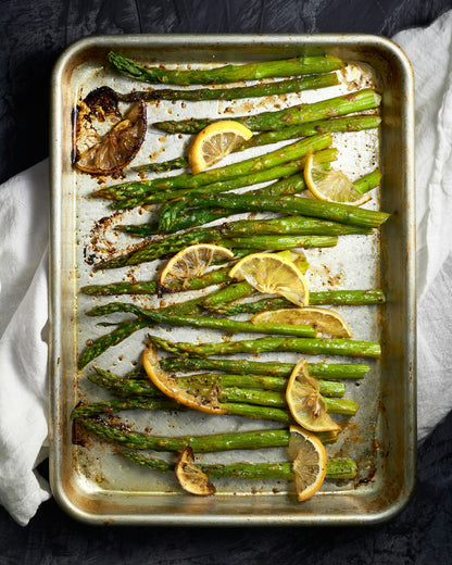 Asparagus, of the variety called, "Mary Washington," previously harvested. There are 22 asparagus spears laying flat within a baking sheet, with lemon slices on top. The asparagus and lemon appear to have already been baked in an oven, and appear grilled.