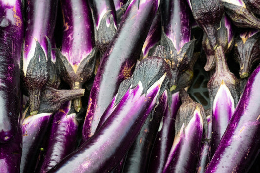 On display are several eggplant, of the variety Long Purple. The eggplant are stacked several layers deep. A small gap between the eggplant shows the underlying black plastic crate containing them. The eggplant have a strikingly purple hue that morphs to a translucent white near the crown, or calyx. The calyx of each eggplant is a dark black color.