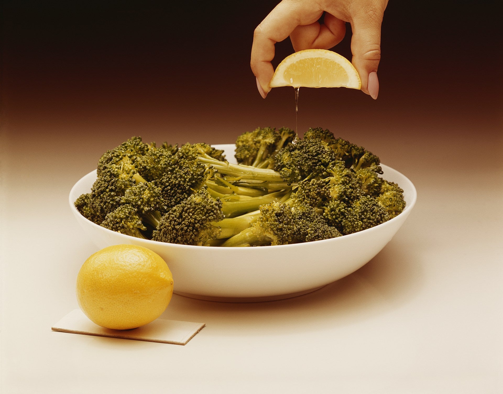 On display is broccoli, of the variety Di Cicco. There is a heaping serving of broccoli spears, laid flat within a white circular serving bowl. The broccoli spears are aligned so their opposing heads gather at the both the right and left sides of the bowl.  In the center, only the spears are visible.  There is an intact lemon resting against the bowl on front left.  A woman's hand is seen descending from the top, squeezing a wedge of lemon juice onto the broccoli.