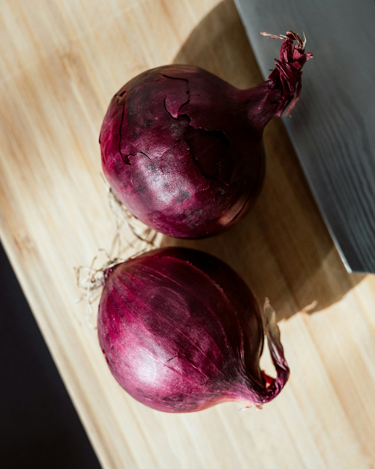 On display are 2 red onions, of the variety Short Red Creole. The red onions display their characteristic dark red shades. The onions are resting on a light wooden butcher block board.