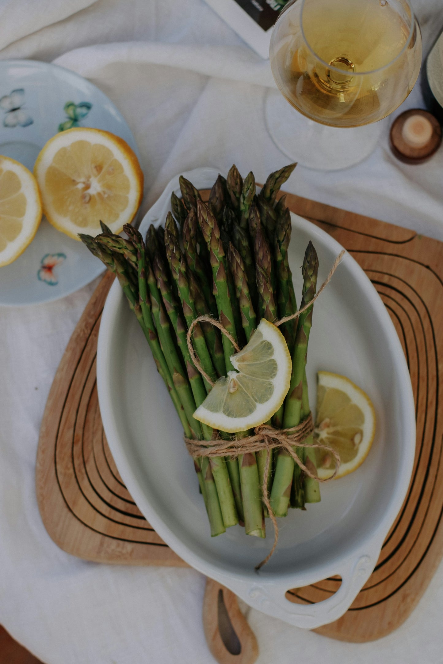 Asparagus, of the variety called, "Mary Washington," previously harvested. There is a collection of asparagus spears, with cooking twine tying them together, within a white porcelain serving dish.  Lemon is adorned on top. 