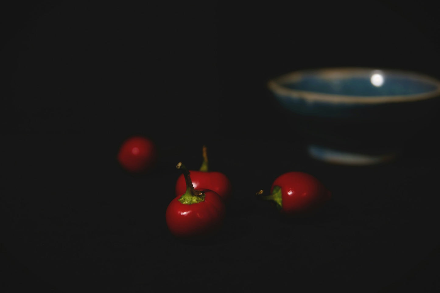 On display are 4 small cherry peppers, of the variety Red Cherry Sweet. Typically known as cherry bombs, these peppers are full of sweetness to enhance any salad. The peppers appear on a dark surface, with 1 pepper prominently in focus in the foreground. There is a slight shadow indicating a light source from the left. In the upper right is an empty blue ceramic bowl. The background is entirely dark, with a low light source, making the entire scene very dark, and full of faint shadows. 